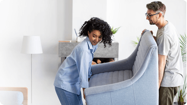 A man and woman carry a large chair through the living room of a home.
